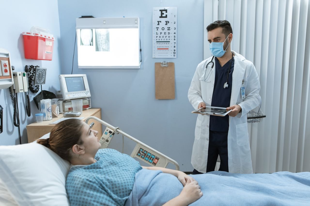 services-01 Doctor wearing face mask consulting with a patient in a hospital room, highlighting healthcare safety.