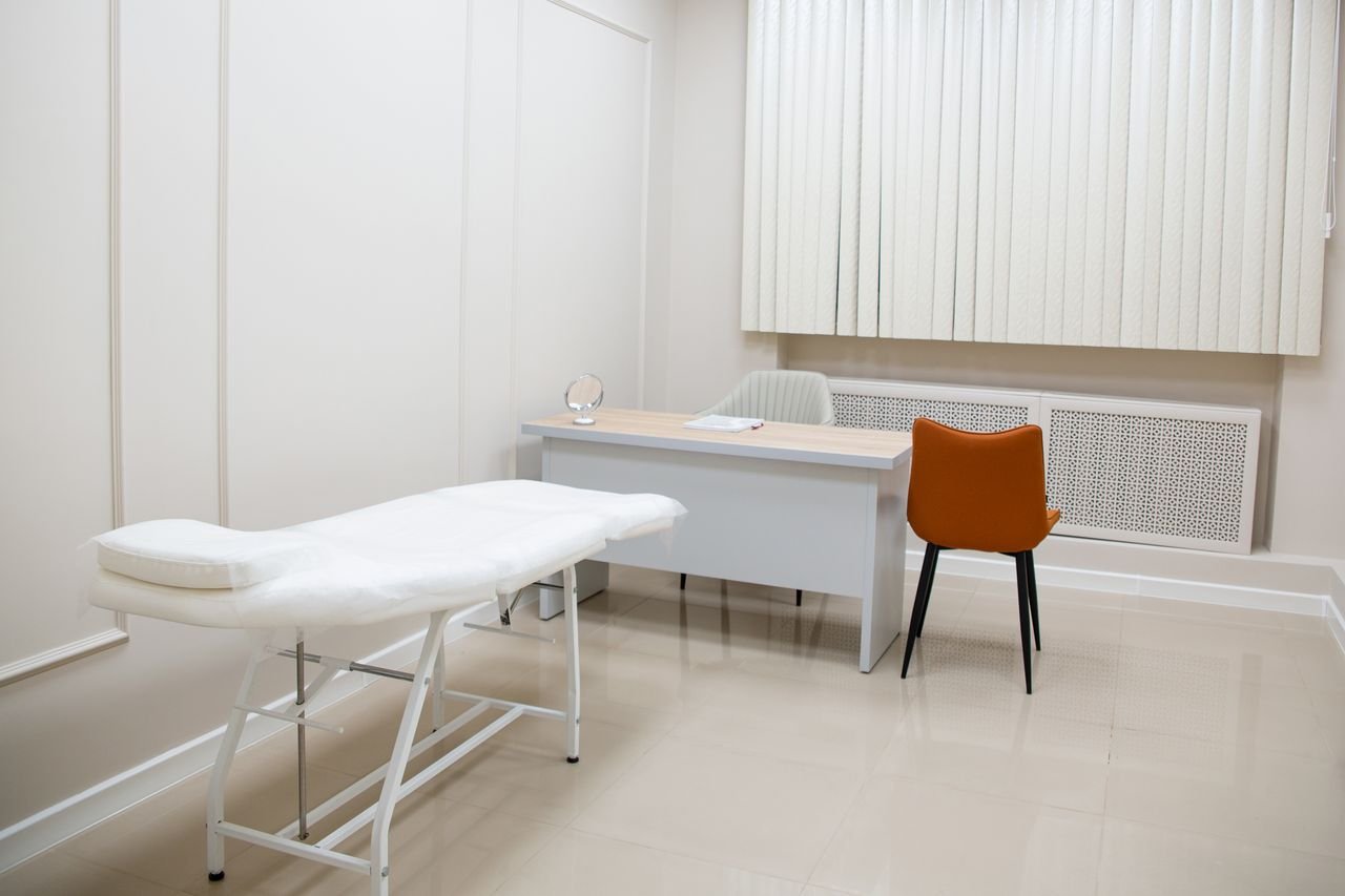 services-01 A minimalist medical examination room featuring a desk, chairs, and an examination bed in a sterile, neutral setting.